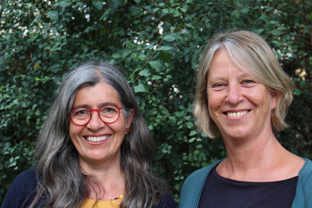 Zwei Frauen mittleren Alters stehen l&auml;chelnd nebeneinander im Freien vor einem gr&uuml;nen, bl&auml;ttrigen Hintergrund.