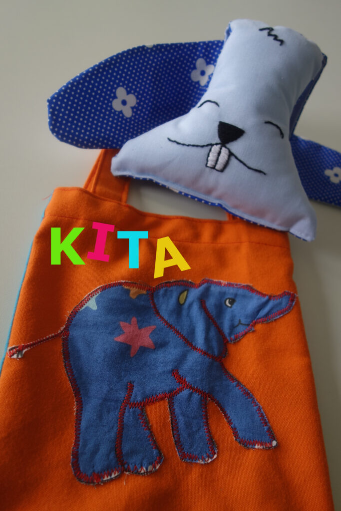 Orange Tasche mit buntem „KITA“-Schriftzug, darauf ein blauer Elefant, oben ein Stofftier mit Hasengesicht und blauen Ohren.